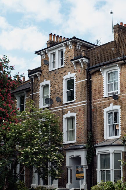 A multi-storey brick terraced house on Lordship Lane in Dulwich, featuring white window frames and decorative architectural details around the windows and roofline. The building has several satellite dishes attached to the brick exterior and a small front garden with trees and shrubs. The scene is set outdoors during the daytime with a partly cloudy sky visible overhead. This image relates to house removals and moving services provided by Removal Company Dulwich, illustrating the type of residential property involved in home relocation and furniture transport processes.