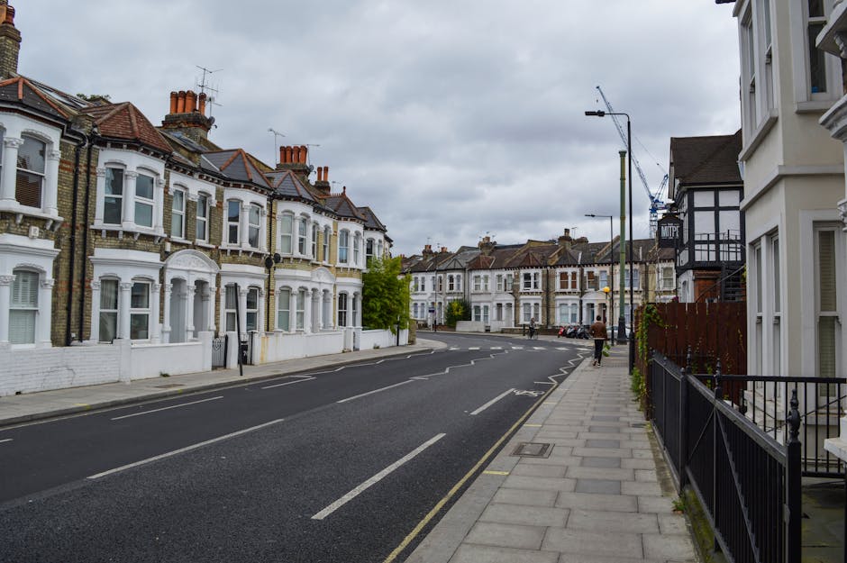 A quiet residential street in Dulwich with rows of Victorian-style terraced houses featuring white facades, bay windows, and red-tiled roofs, under an overcast sky. The pavement on the right side has a black iron fence and a narrow walkway, with a person walking away from the camera. The street is lined with parked cars and is equipped with streetlights and a crane in the distance, suggesting ongoing building or maintenance work. The scene captures a typical home environment where removals and furniture transport might take place, with potential access points for house removals services by Removal Company Dulwich, aligned with house relocation or packing and moving processes.