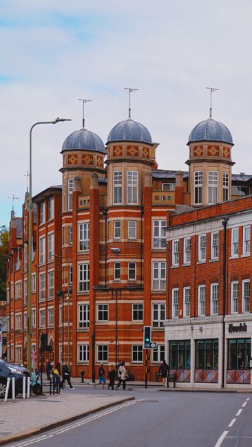 The image shows a multi-storey red brick building with distinctive turreted towers topped with conical grey roofs, situated at a street corner. The building features large windows with white frames an
